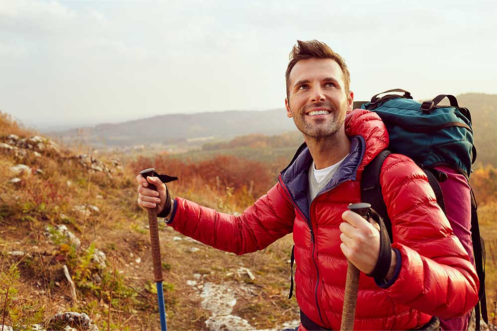 A man in a red puffer coat smiles on a trail holding hiking poles.